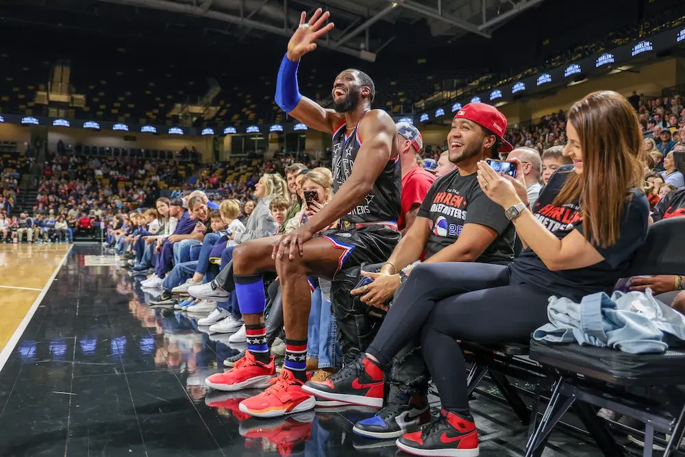 A Harlem Globetrotters player sitting on the lap of a fan in the front row waving to the crowd.